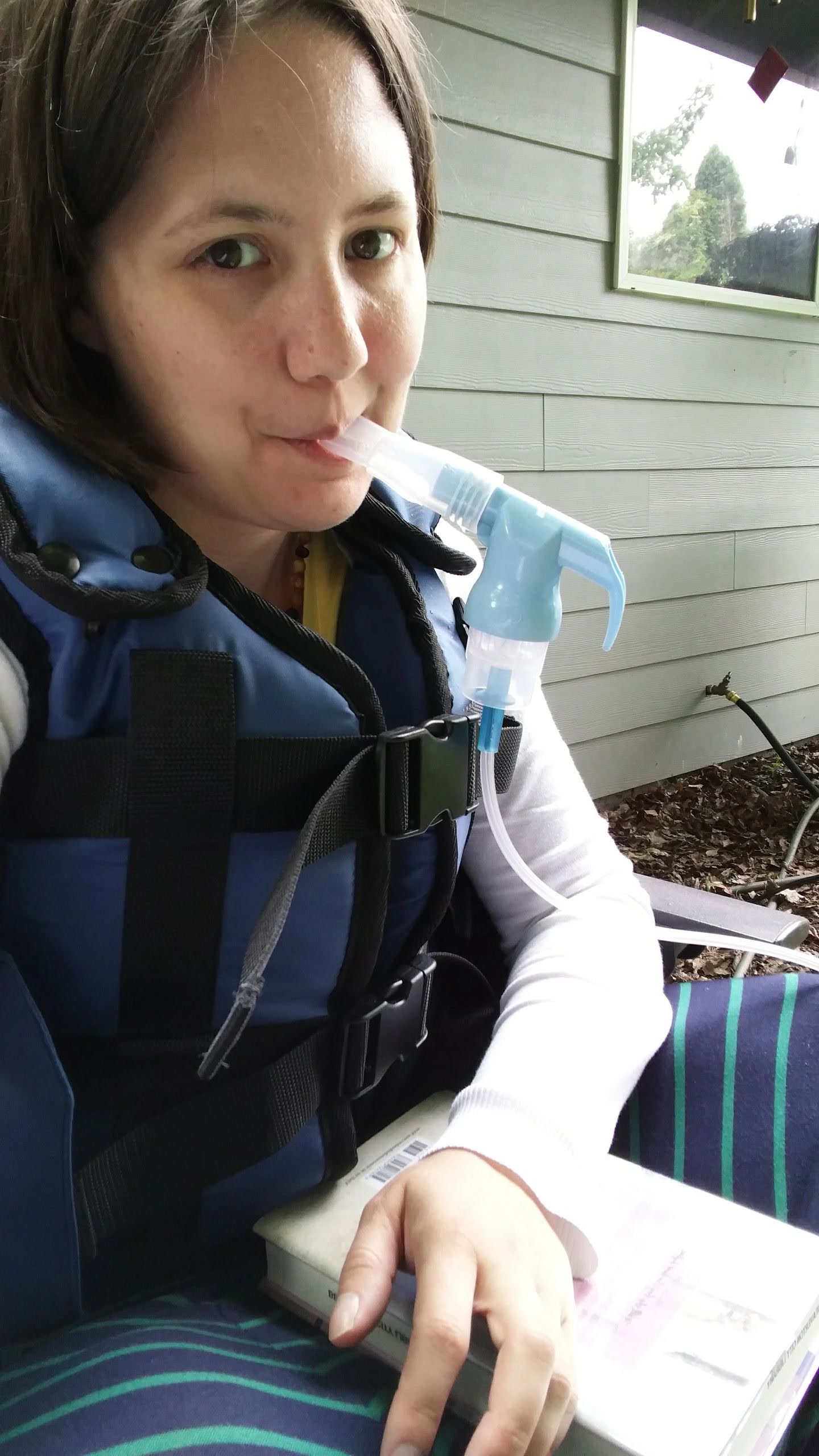 Cindy Baldwin wearing an airway-clearance vest and using a nebulizer while reading a book on a porch